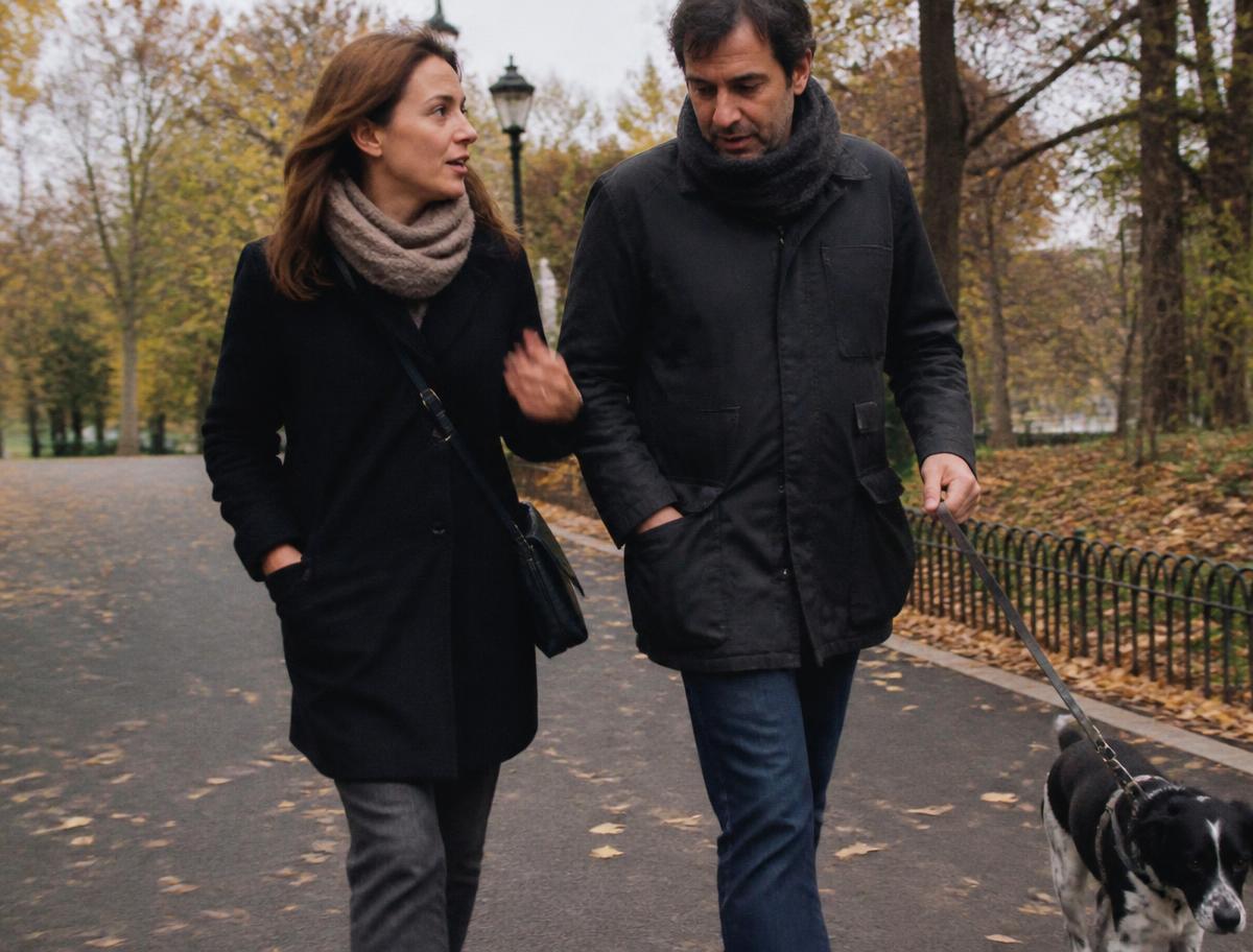 Claudia y Sergio paseando al perro por un parque en otoño, hablando entre ellos mientras caminan