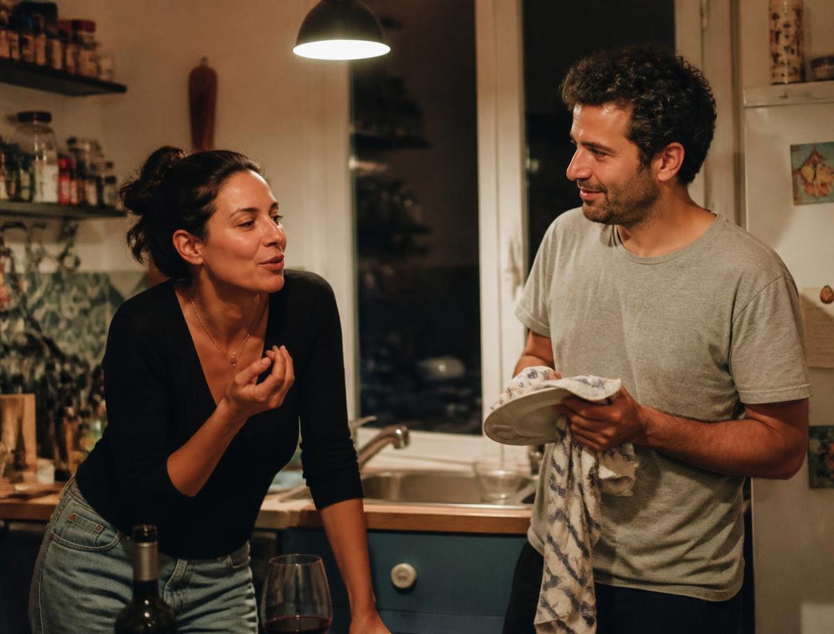 Sofía y Daniel conversando en la cocina de casa por la noche; ella apoyada en la encimera y él secando un plato