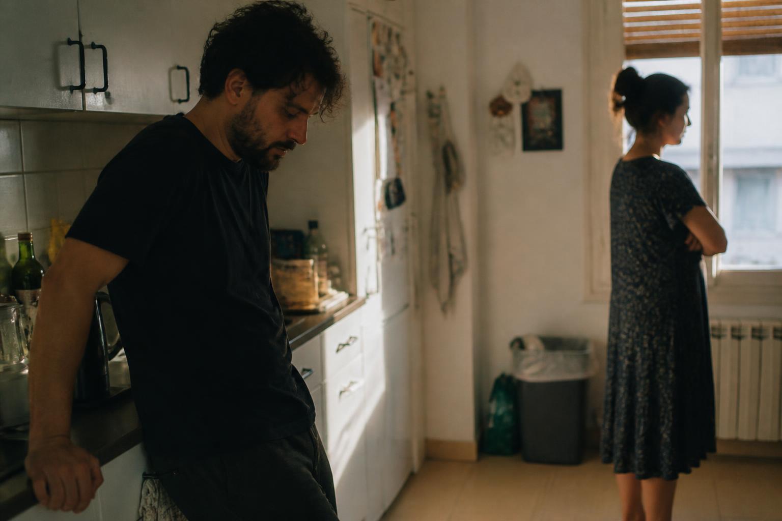 Él apoyado en la encimera mirando al suelo y ella junto a la ventana con los brazos cruzados, tras una discusión en la cocina