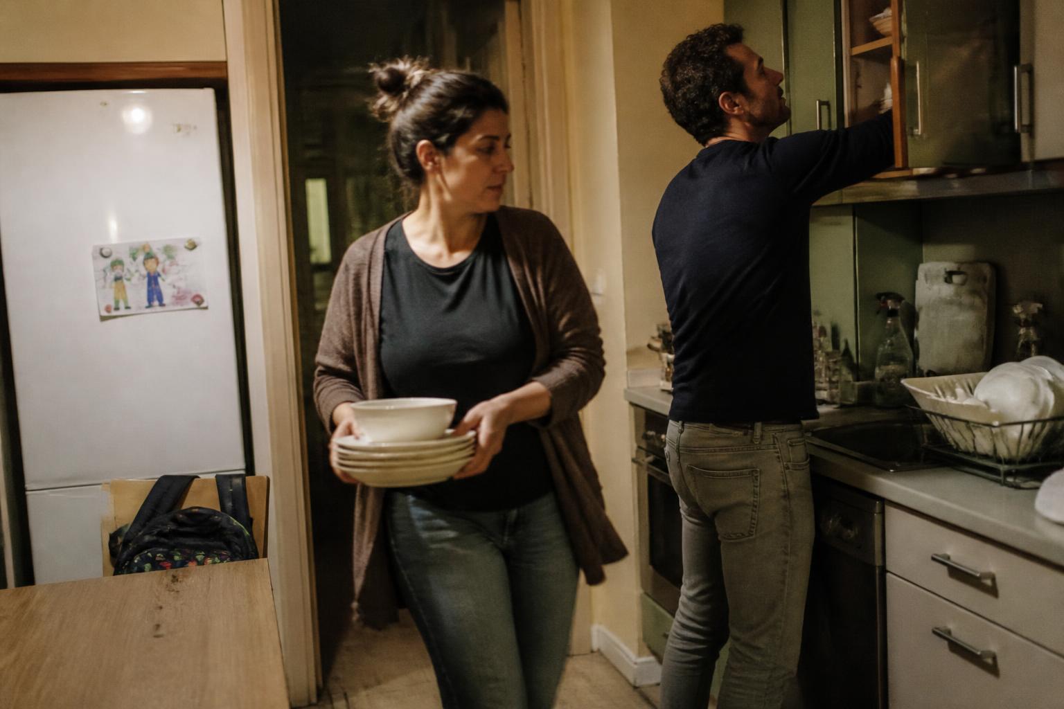 Pareja recogiendo la cocina de noche: ella lleva platos y él abre un armario, con la mochila del niño y un dibujo en la nevera